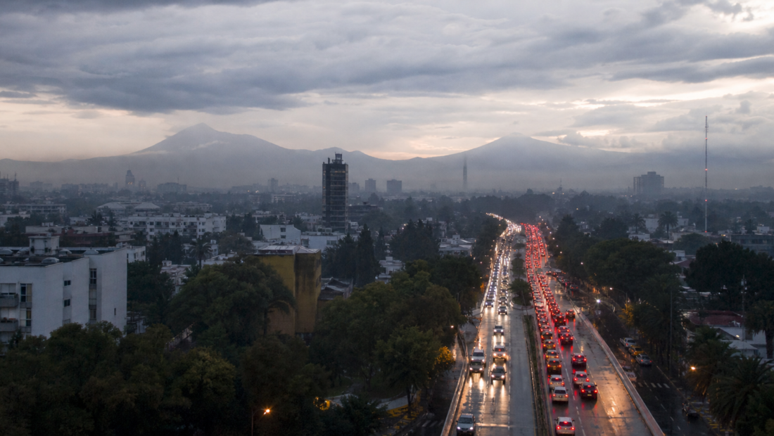 Tráfico vehicular en una avenida de la Ciudad de México al amanecer, con pavimento mojado y cielo nublado.